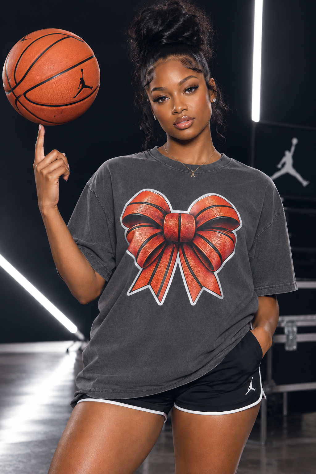 A woman posing in a dark gray t-shirt with a large red and orange basketball bow graphic on the front, holding a basketball on her finger. The background is a dark studio setting with neon light strips and a faint Jordan logo.
